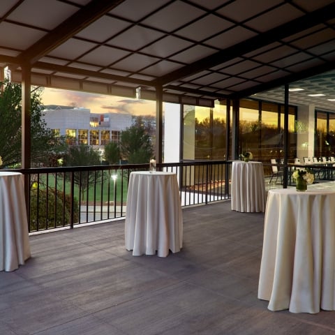 White draped high-top tables stand on a covered outdoor patio overlooking a building and trees at sunset, adjacent to an indoor event space with glass walls.