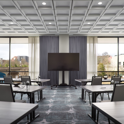 A large monitor stands centered in a modern conference room with tables and chairs set for meetings, overlooking a city. Water bottles read "THE TENNESSEE".