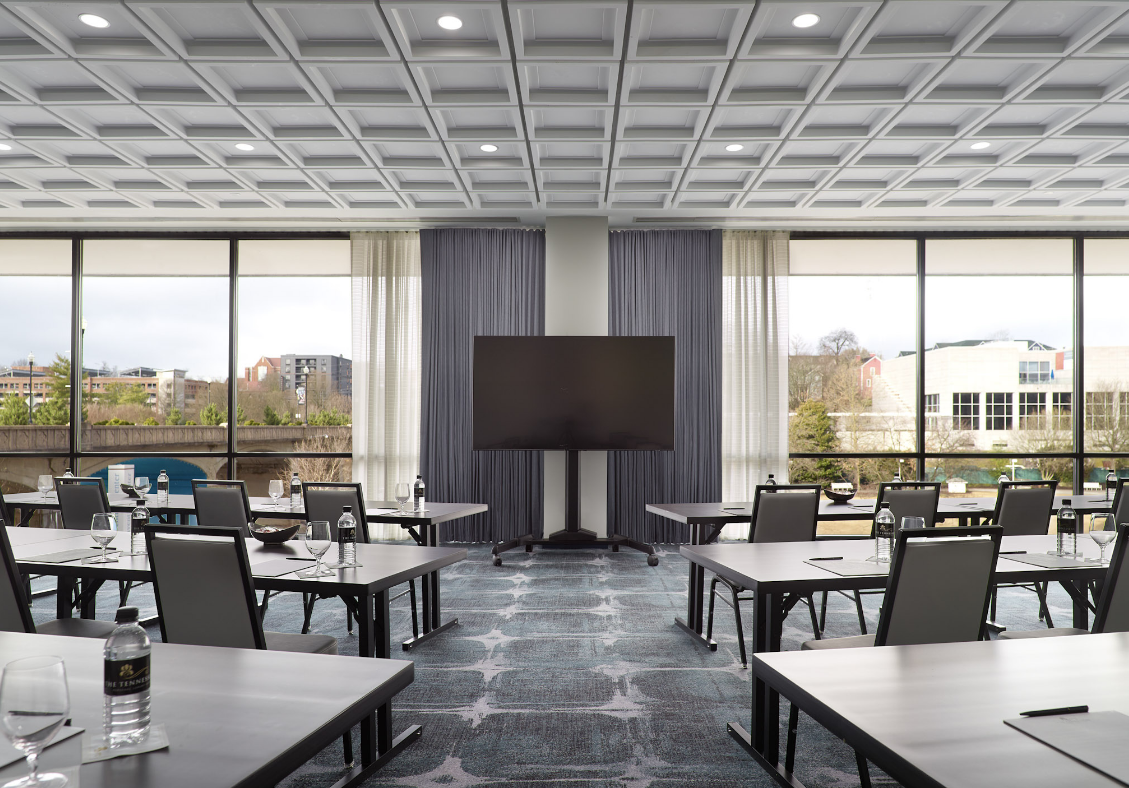 A large monitor stands centered in a modern conference room with tables and chairs set for meetings, overlooking a city. Water bottles read "THE TENNESSEE".