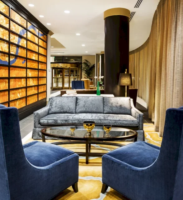 A grey sofa and two blue armchairs face a glass coffee table in a modern lounge. Features include a textured orange wall art, dark pillar, and golden curtains on a yellow rug.