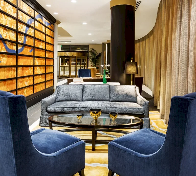 A grey sofa and two blue armchairs face a glass coffee table in a modern lounge. Features include a textured orange wall art, dark pillar, and golden curtains on a yellow rug.