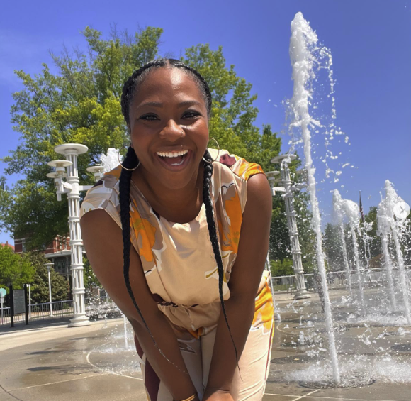 A woman with braids laughs joyfully, bending near spraying water jets in an outdoor fountain on a sunny day with green trees in the background.