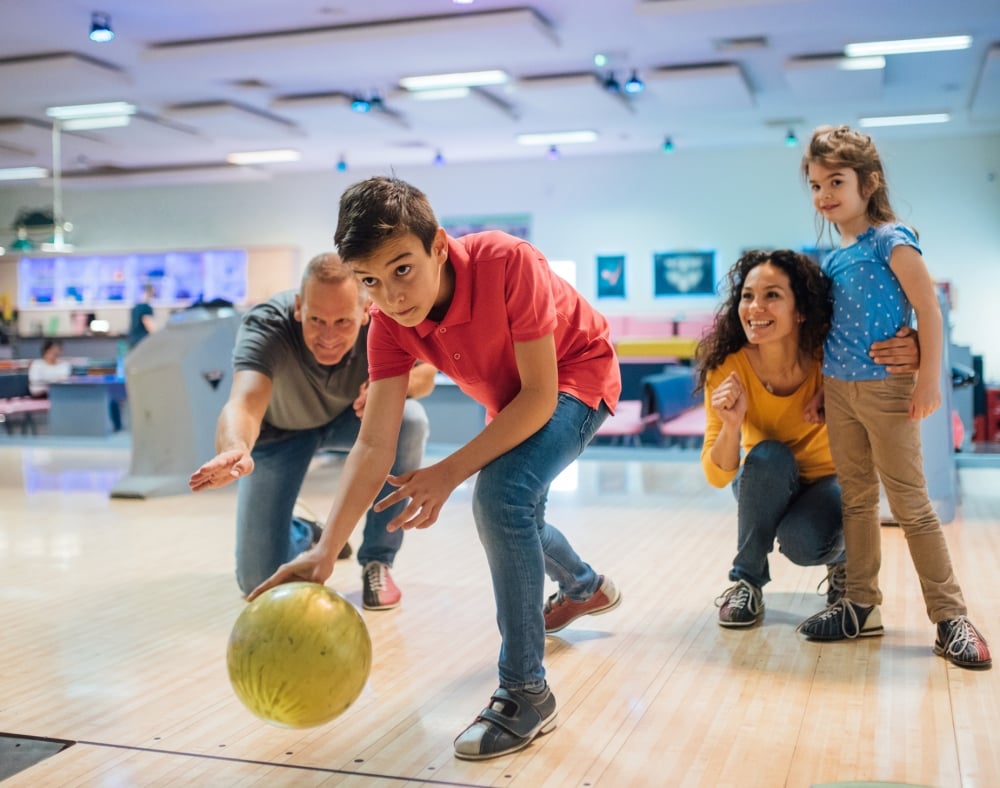 A young boy rolls a yellow bowling ball down a lane, watched by an encouraging man and a smiling woman holding a small girl, inside a bowling alley.