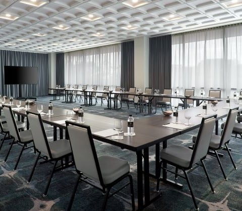 A U-shaped conference table arrangement with chairs, water bottles, and glasses is set for a meeting in a spacious room featuring a textured ceiling, patterned carpet, and large windows.