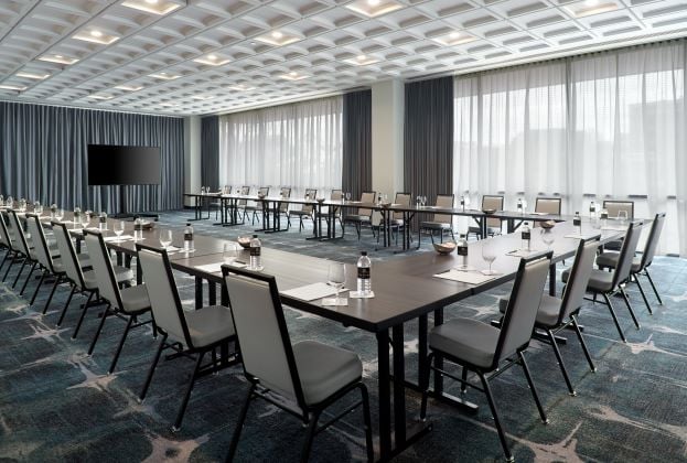 A U-shaped conference table arrangement with chairs, water bottles, and glasses is set for a meeting in a spacious room featuring a textured ceiling, patterned carpet, and large windows.
