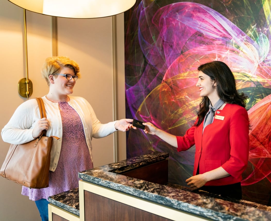A smiling guest receives a key card from a hotel staff member at a marbled reception desk with a vibrant abstract mural. Text: Hilton Honors (on card and name tag), Hilton (on name tag).