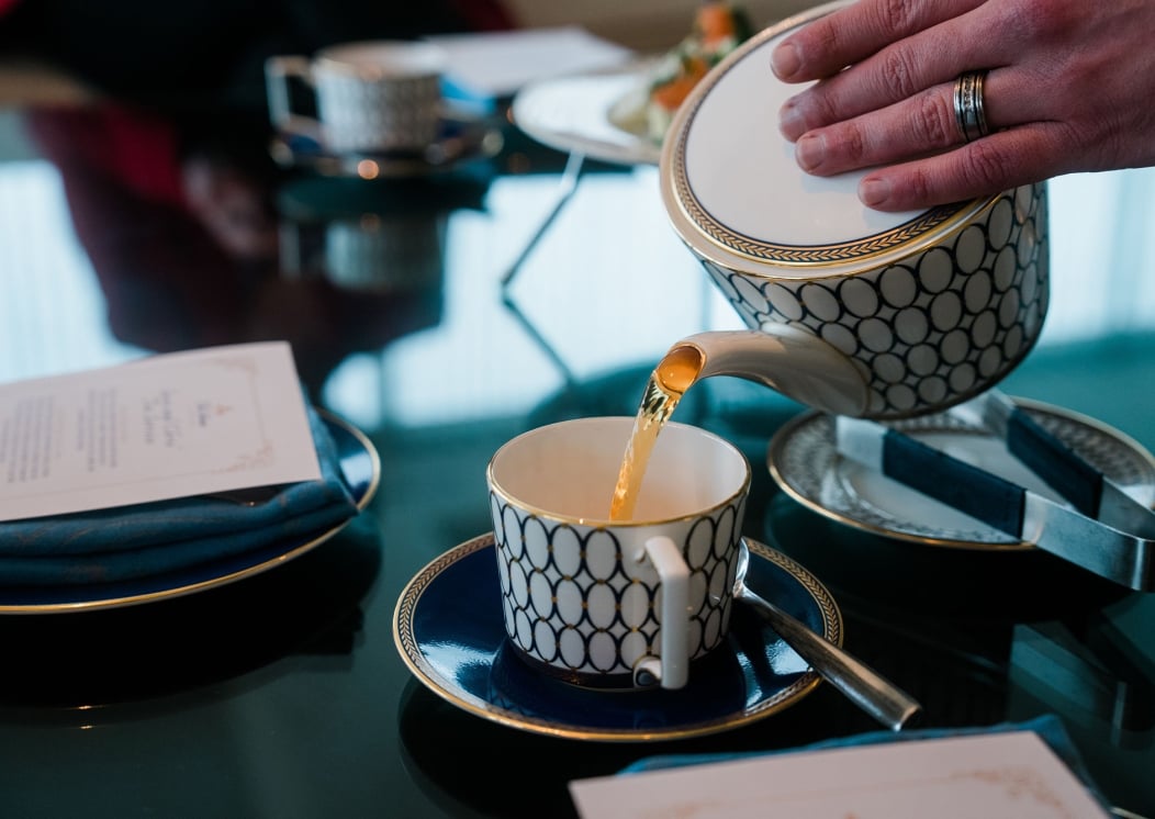 A hand pours golden tea from a white and navy patterned teapot into a matching teacup, sitting on a dark, reflective table alongside a menu, saucer, and sugar tongs.