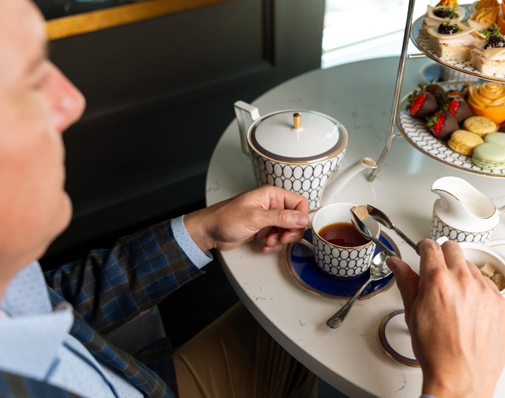 A man's hands add a sugar cube to his teacup. He sits at a white table with a patterned teapot, milk jug, sugar bowl, and a tiered stand displaying sweets and savories.