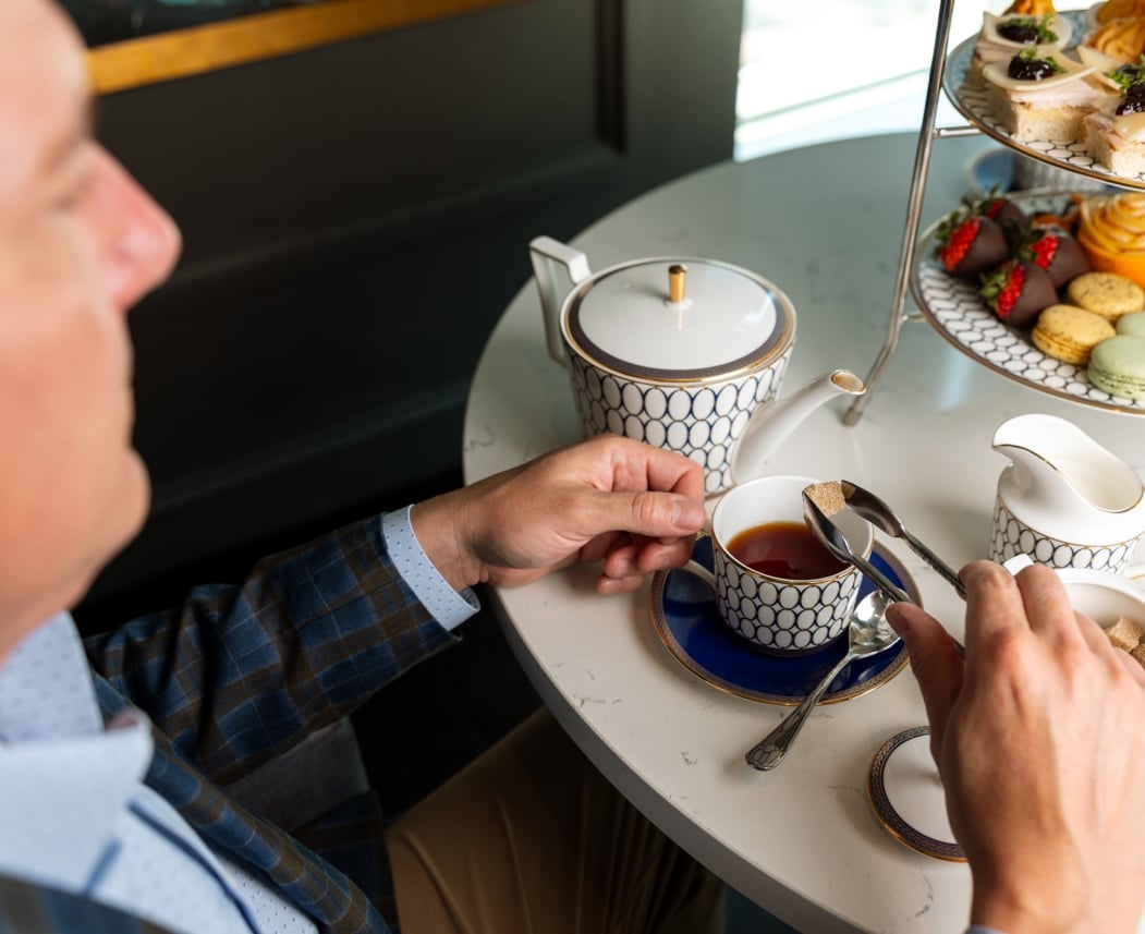 A man's hands add a sugar cube to his teacup. He sits at a white table with a patterned teapot, milk jug, sugar bowl, and a tiered stand displaying sweets and savories.