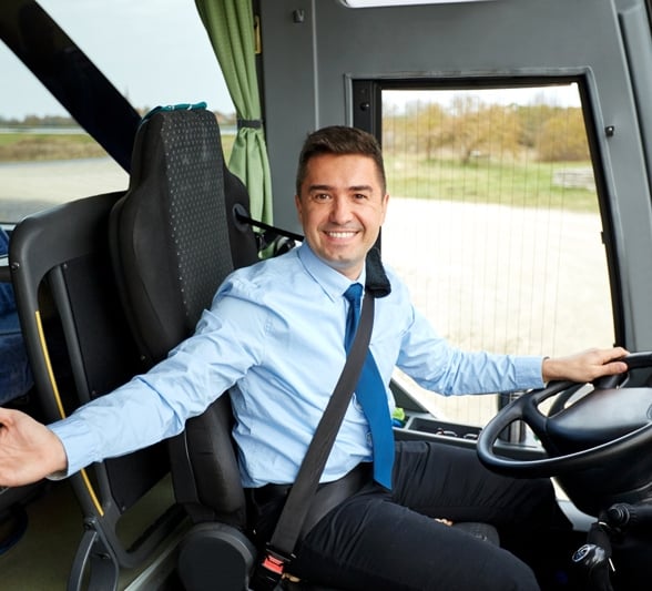 A smiling bus driver in a blue shirt and tie sits at the wheel, gesturing with one hand, inside a bus with passenger seats and an outdoor view.