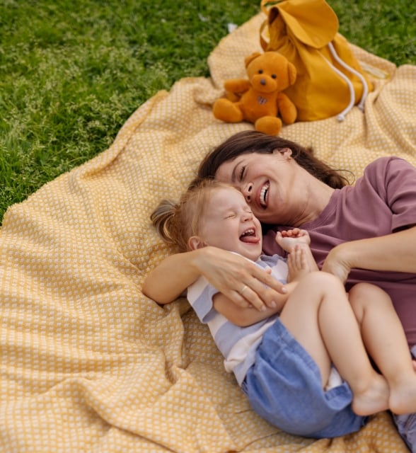 A woman and toddler lie embracing and laughing on a yellow blanket over green grass, with a yellow backpack and teddy bear nearby.