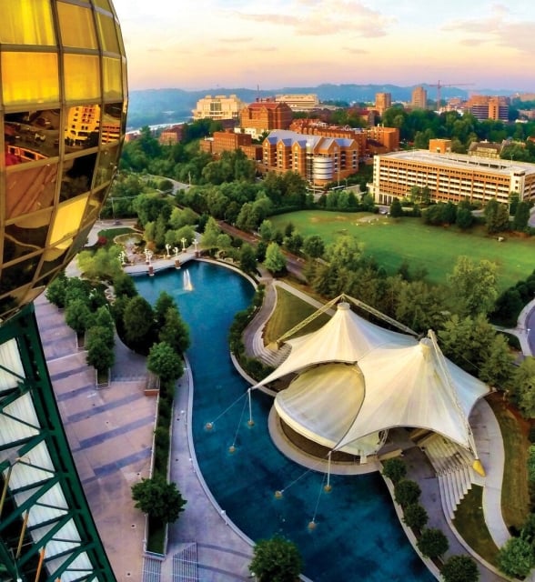 A large golden sphere stands overlooking an urban park featuring a winding blue pond, a white tented pavilion, green spaces, and a cityscape against a sunset sky.