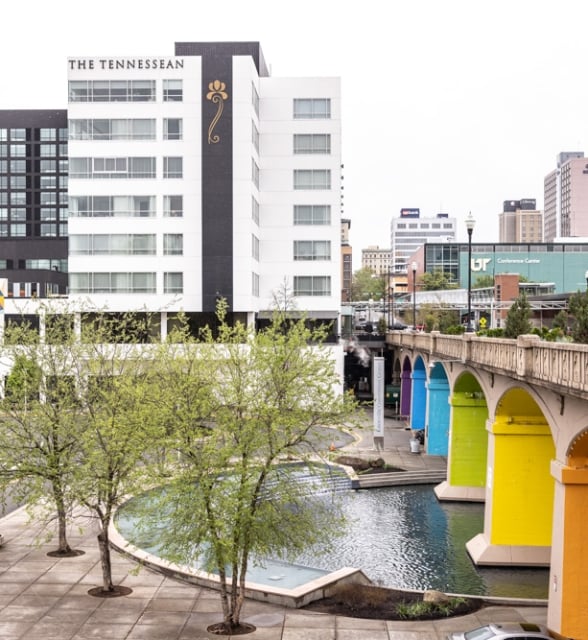 THE TENNESSEAN and "EXCHANGE" hotels stand by a stone bridge with rainbow-colored arches spanning a canal. An urban cityscape with trees and a "Knoxville Convention Center" sign.
