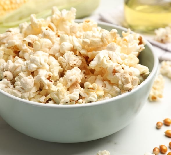 Fluffy white popcorn fills a light blue bowl. It sits on a white surface amidst scattered kernels, a corn cob, and cooking oil in the background.