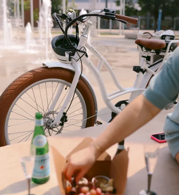 A white "PRIORITY" bicycle with brown tires is parked near a fountain. A person reaches into a food box next to a "S.PELLEGRINO" bottle.
