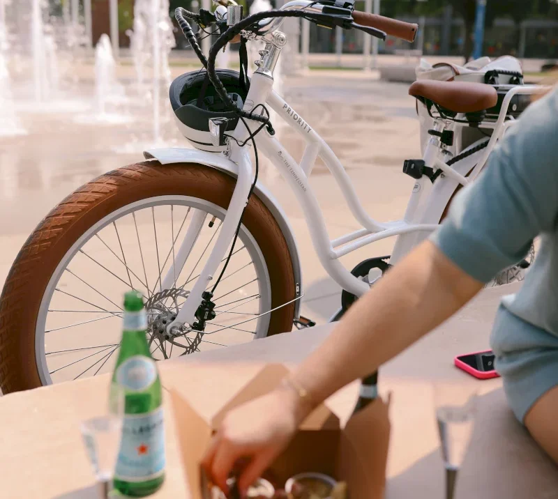 A white "PRIORITY" bicycle with brown tires is parked near a fountain. A person reaches into a food box next to a "S.PELLEGRINO" bottle.