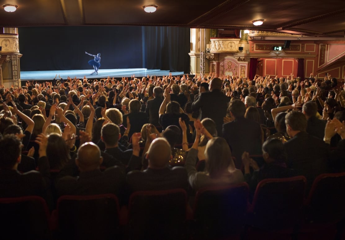 A ballerina performs on stage as a large, applauding audience fills a dimly lit theater. An **EXIT** sign with an arrow is visible above the seating area.
