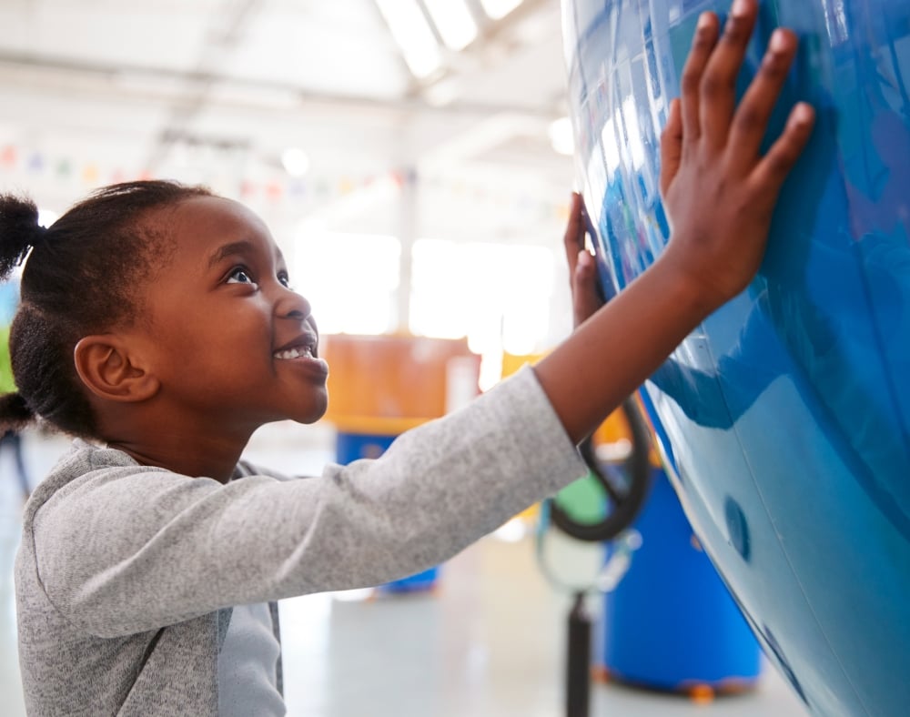 A smiling young Black girl looks up, touching a large, curved blue object with both hands in a bright, spacious building, likely a museum or science center.