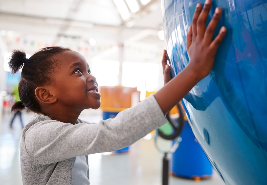A smiling young Black girl looks up, touching a large, curved blue object with both hands in a bright, spacious building, likely a museum or science center.