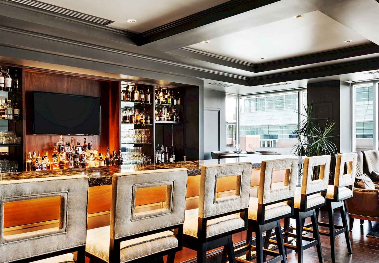 Elegant bar stools line a dark marble bar counter displaying many spirit bottles and glasses. This upscale lounge features large windows overlooking city buildings.