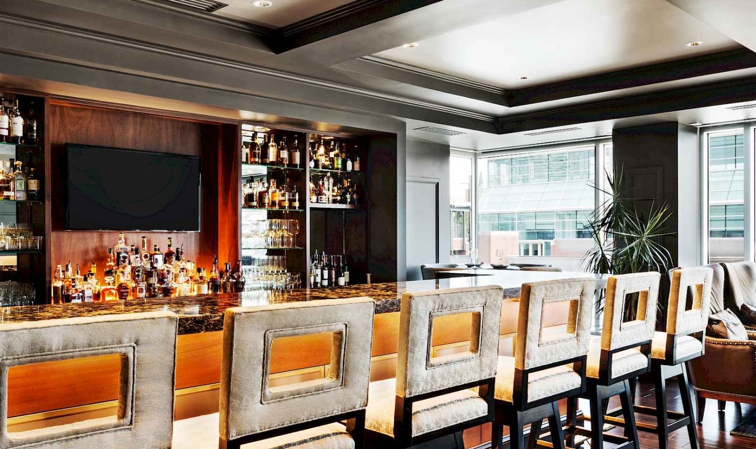 Elegant bar stools line a dark marble bar counter displaying many spirit bottles and glasses. This upscale lounge features large windows overlooking city buildings.