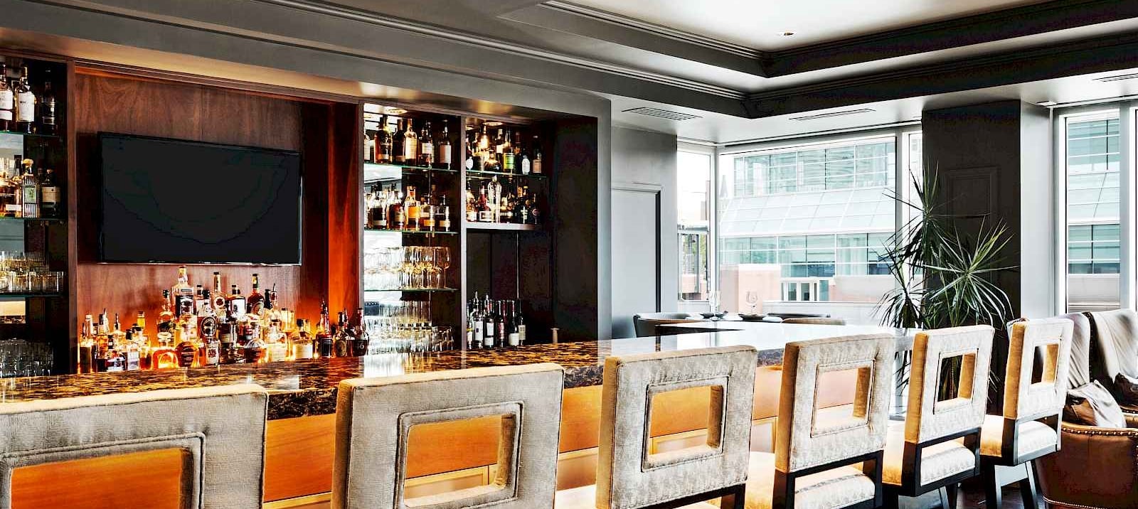 Elegant bar stools line a dark marble bar counter displaying many spirit bottles and glasses. This upscale lounge features large windows overlooking city buildings.