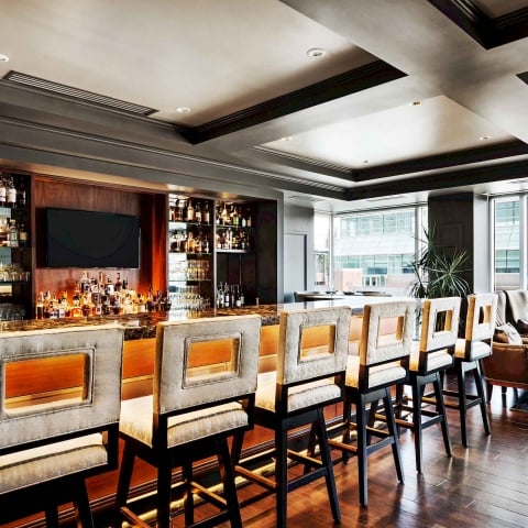 Elegant bar stools line a dark marble bar counter displaying many spirit bottles and glasses. This upscale lounge features large windows overlooking city buildings.