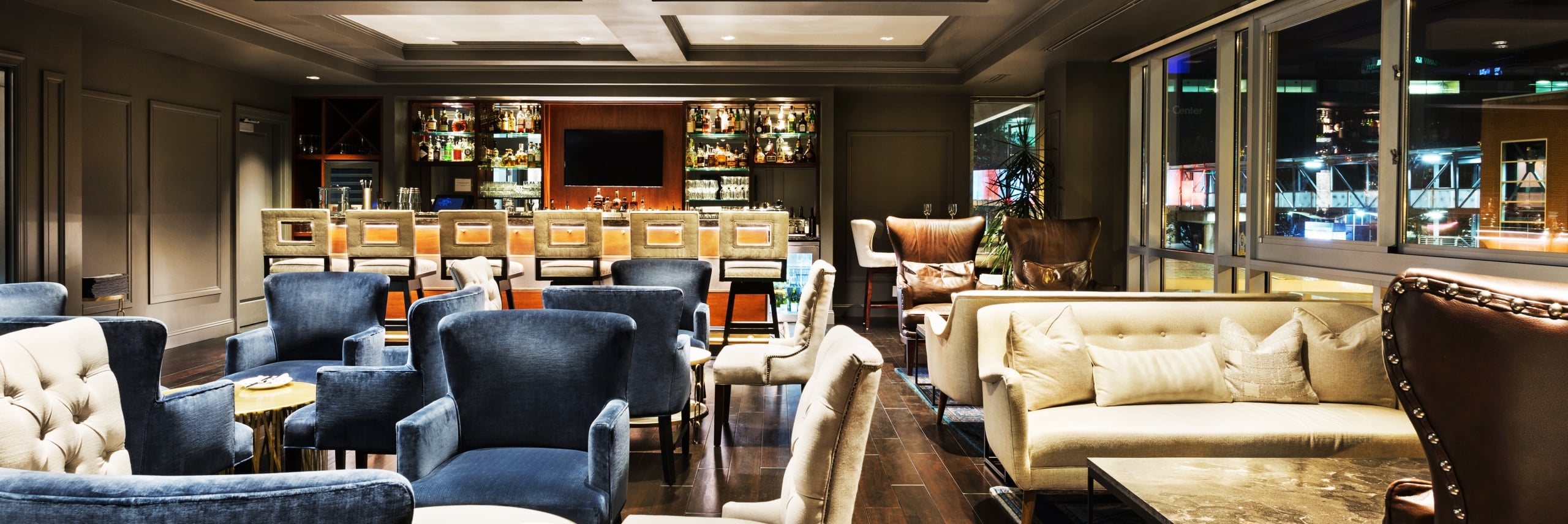 A luxurious hotel lounge featuring plush blue armchairs, a cream sofa, dark wood floors, and a well-lit bar area.