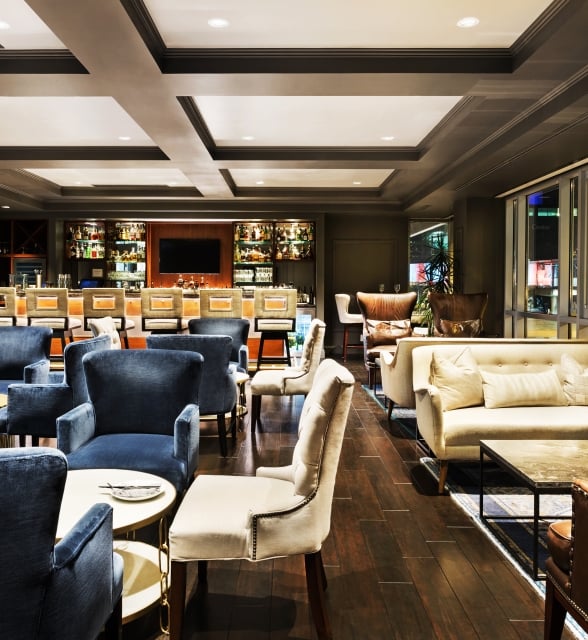 A luxurious hotel lounge featuring plush blue armchairs, a cream sofa, dark wood floors, and a well-lit bar area.