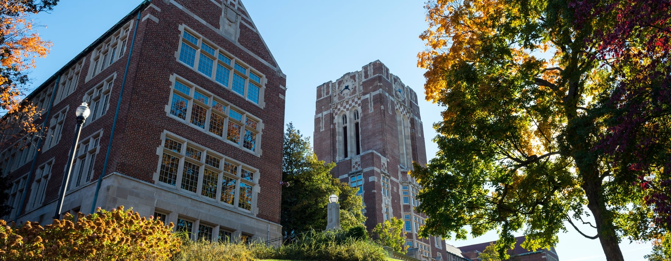 Two large brick university buildings, one with a prominent clock tower, overlook a grassy hill, framed by vibrant autumn trees under a bright blue sky.