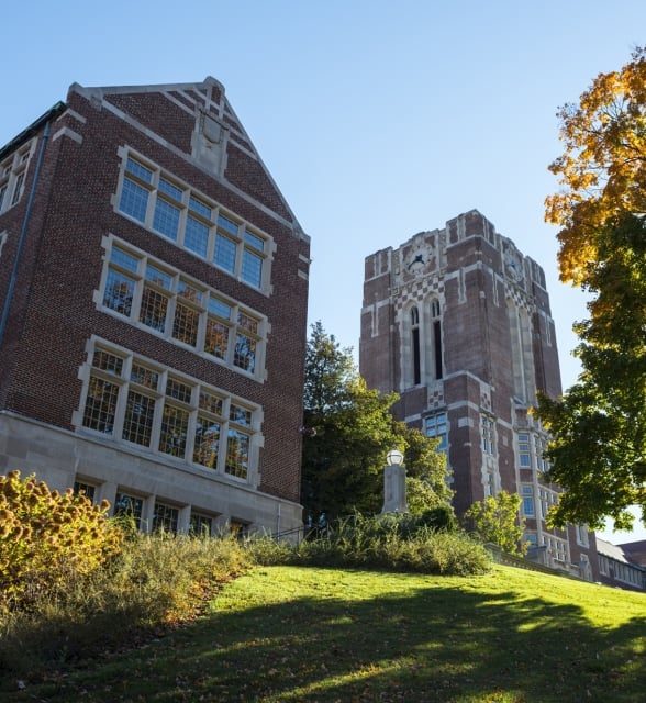 A gabled brick building and a clock tower stand on a sunlit grassy hill with autumn foliage under a clear blue sky.