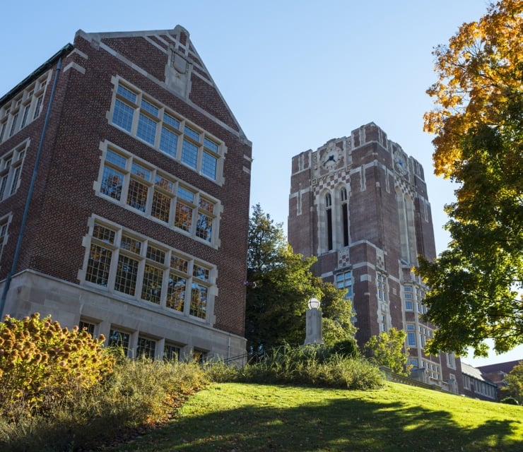 A gabled brick building and a clock tower stand on a sunlit grassy hill with autumn foliage under a clear blue sky.