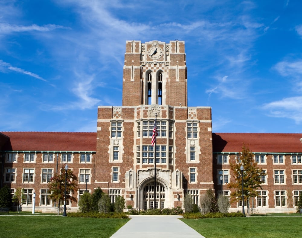 A grand brick university building with a central clock tower stands on a green lawn. An American flag flies above the entrance, under a bright blue sky with wispy clouds.