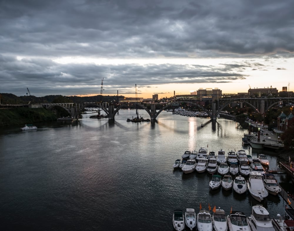 A wide river flows under multiple arched bridges, past an urban landscape with many docked boats, under a cloudy twilight sky.