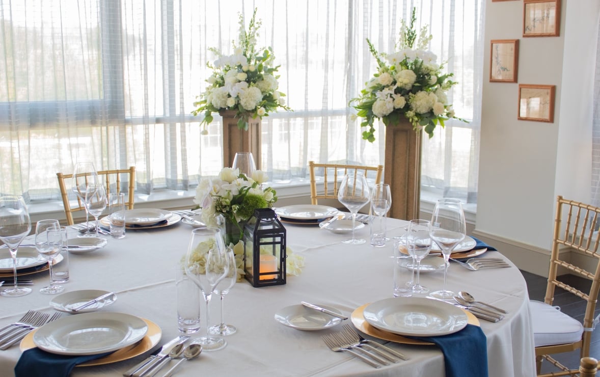 A round dining table is formally set with place settings, a floral centerpiece, and a lantern. It stands in a bright room with large windows and tall white floral arrangements.
