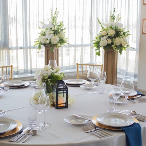 A round dining table is formally set with place settings, a floral centerpiece, and a lantern. It stands in a bright room with large windows and tall white floral arrangements.