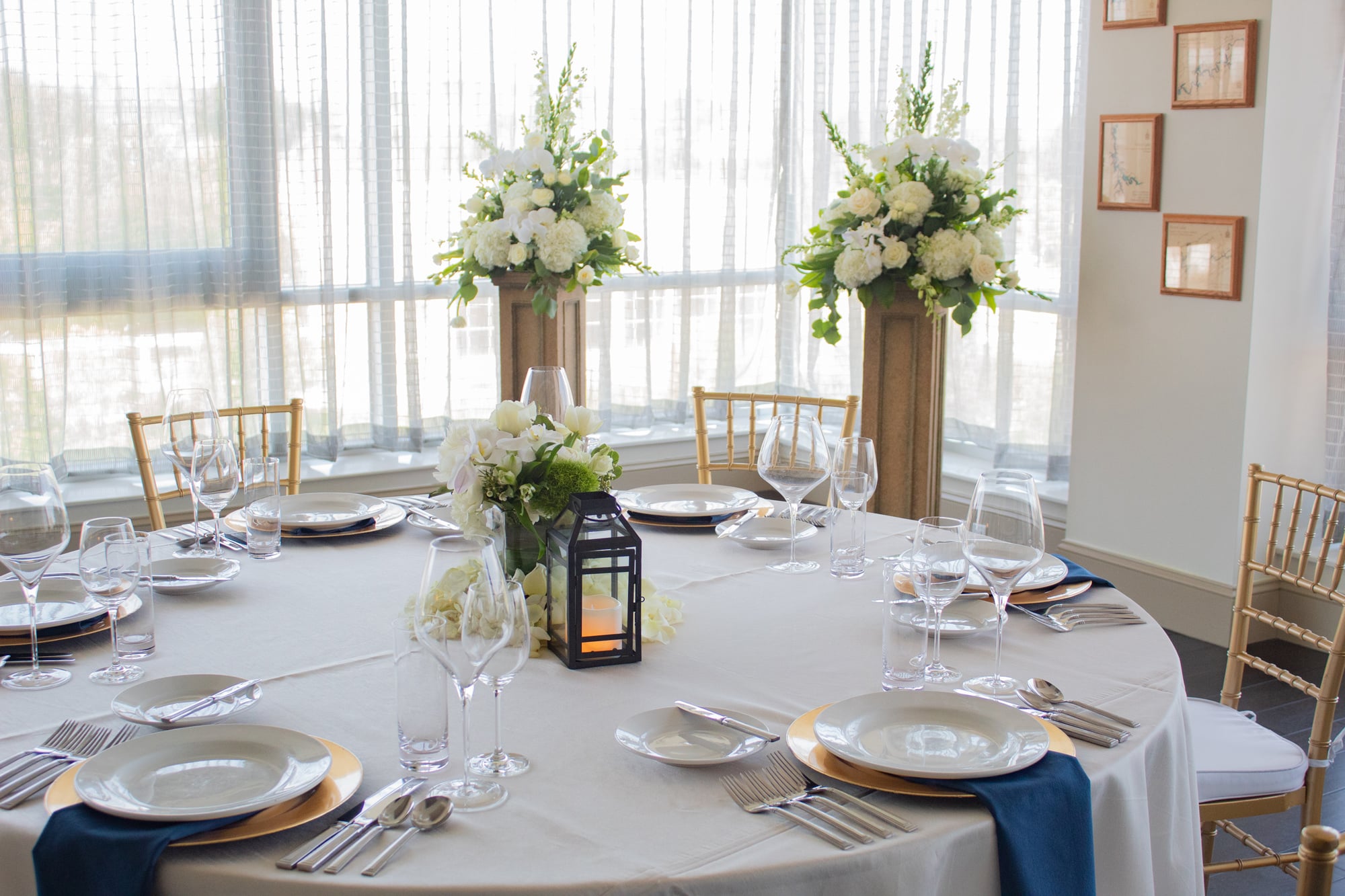 A round dining table is formally set with place settings, a floral centerpiece, and a lantern. It stands in a bright room with large windows and tall white floral arrangements.