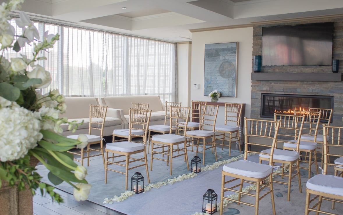 Gold chairs with white cushions are arranged in rows on a blue aisle lined with petals and lanterns, facing a fireplace and large windows in a bright room.
