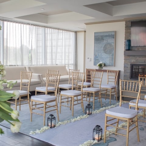 Gold chairs with white cushions are arranged in rows on a blue aisle lined with petals and lanterns, facing a fireplace and large windows in a bright room.