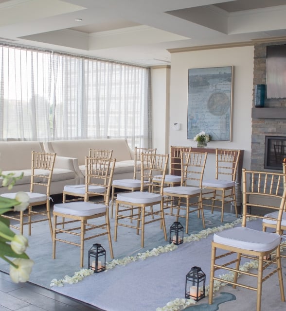 Gold chairs with white cushions are arranged in rows on a blue aisle lined with petals and lanterns, facing a fireplace and large windows in a bright room.