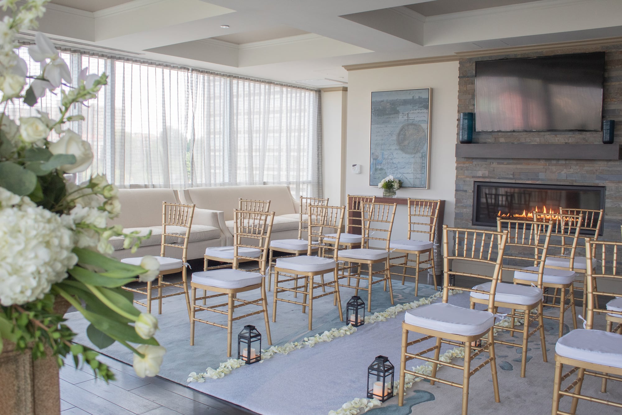 Gold chairs with white cushions are arranged in rows on a blue aisle lined with petals and lanterns, facing a fireplace and large windows in a bright room.