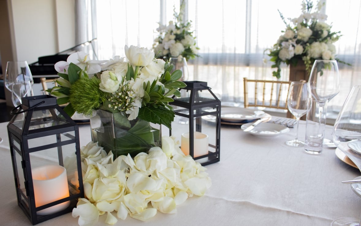 A white and green floral centerpiece rests on a white tablecloth, flanked by black lanterns with LED candles and surrounded by rose petals, part of a formal table setting.