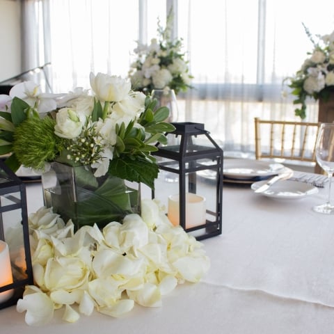 A white and green floral centerpiece rests on a white tablecloth, flanked by black lanterns with LED candles and surrounded by rose petals, part of a formal table setting.
