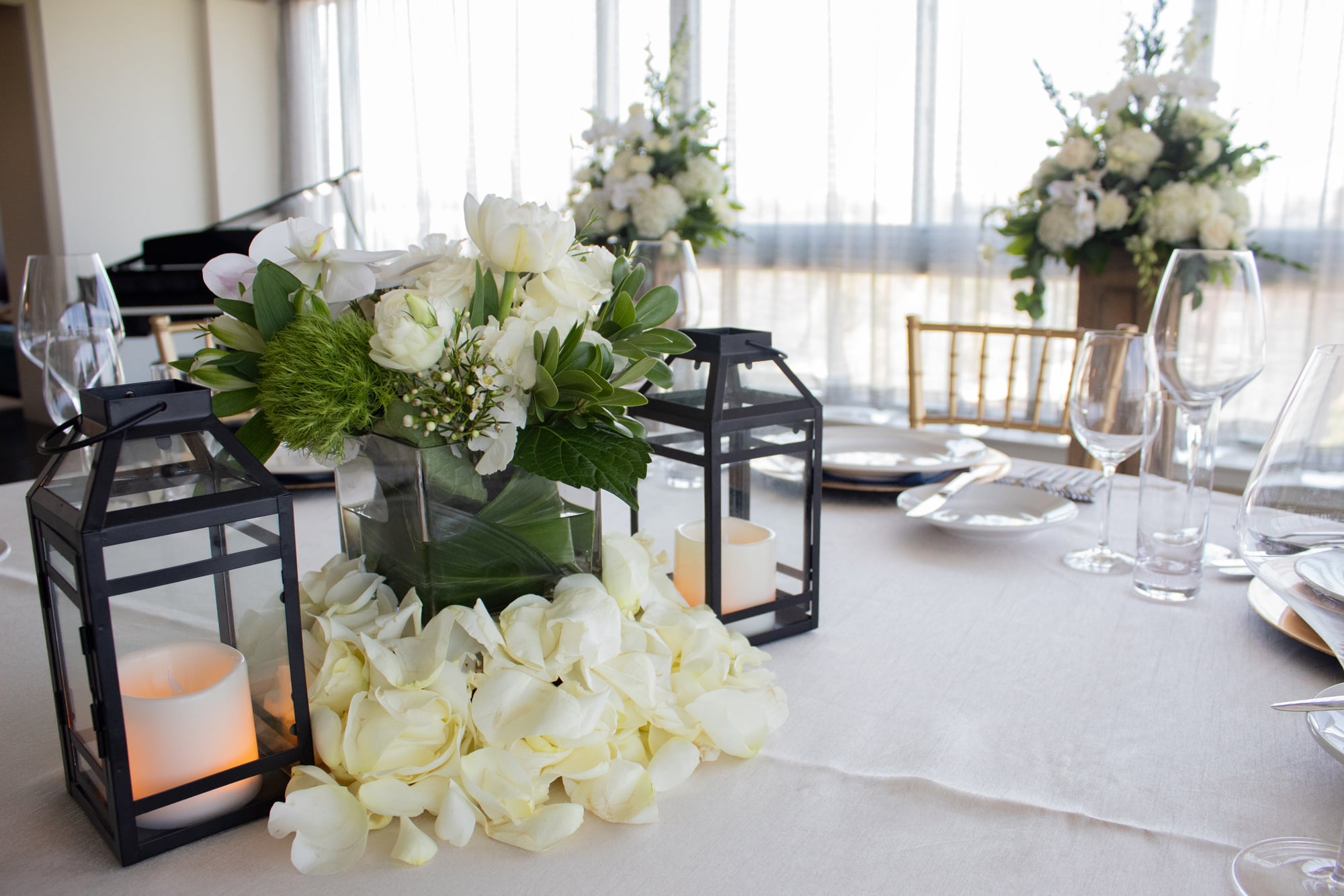 A white and green floral centerpiece rests on a white tablecloth, flanked by black lanterns with LED candles and surrounded by rose petals, part of a formal table setting.