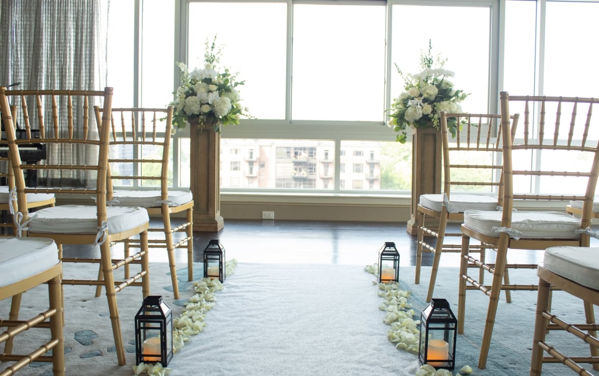 A wedding aisle, lined with white rose petals and lanterns, flanked by gold chairs, leads to two white floral arrangements on pedestals before bright windows overlooking a city.