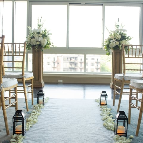 A wedding aisle, lined with white rose petals and lanterns, flanked by gold chairs, leads to two white floral arrangements on pedestals before bright windows overlooking a city.