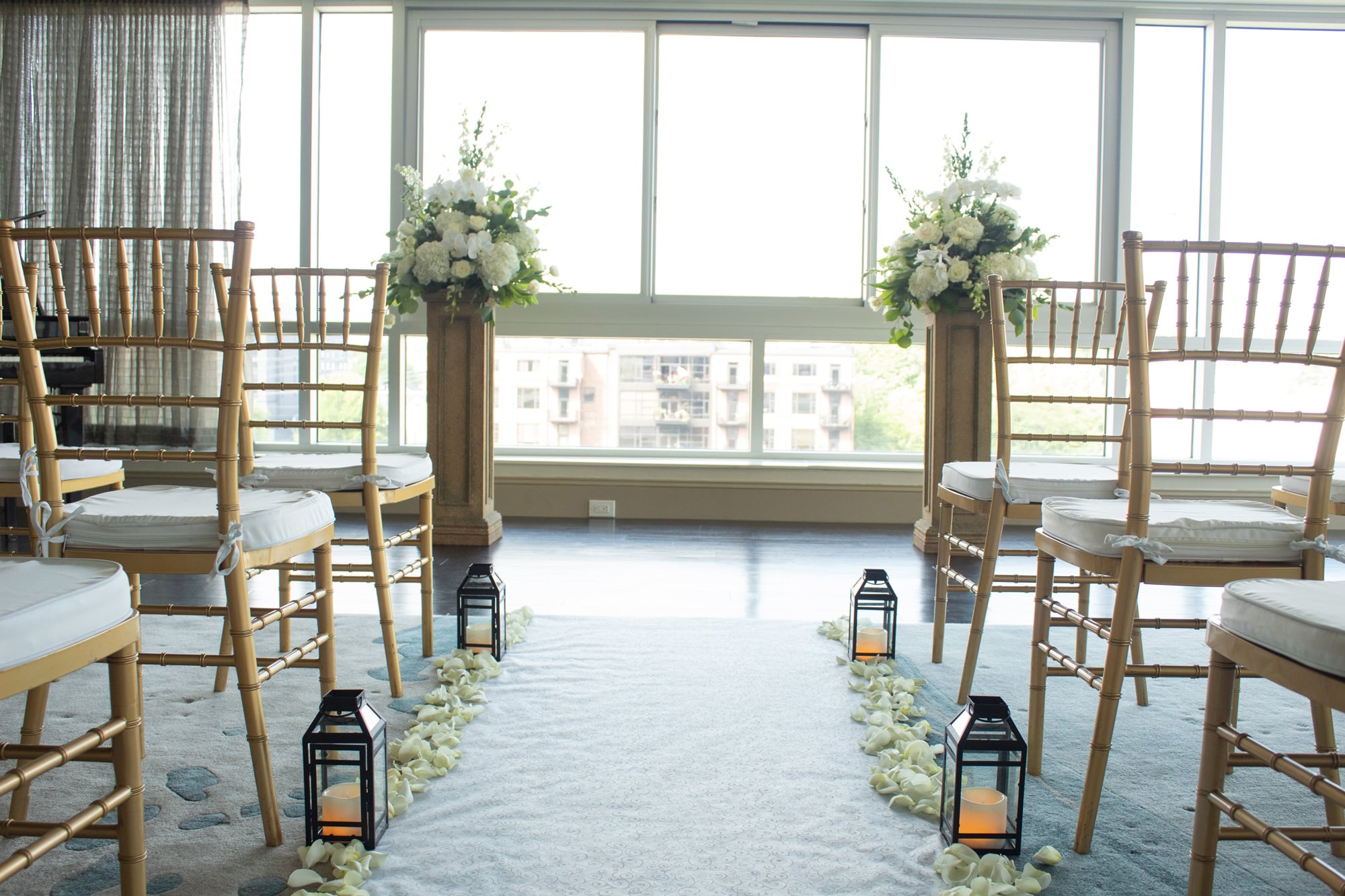 A wedding aisle, lined with white rose petals and lanterns, flanked by gold chairs, leads to two white floral arrangements on pedestals before bright windows overlooking a city.