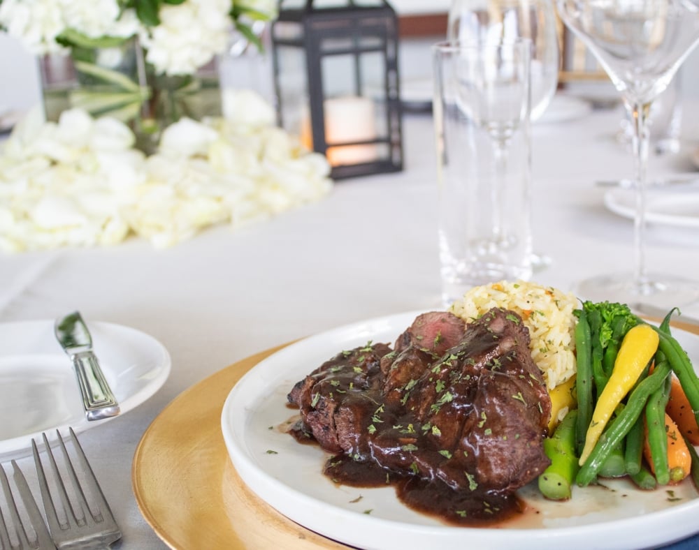 Steak with sauce, rice, and colorful steamed vegetables are plated on a white dish, on a wooden charger, at a formal dining table with white flowers and glassware.
