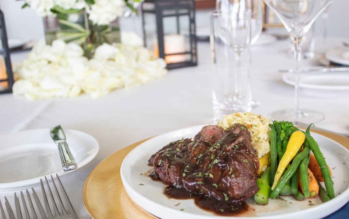 Steak with sauce, rice, and colorful steamed vegetables are plated on a white dish, on a wooden charger, at a formal dining table with white flowers and glassware.