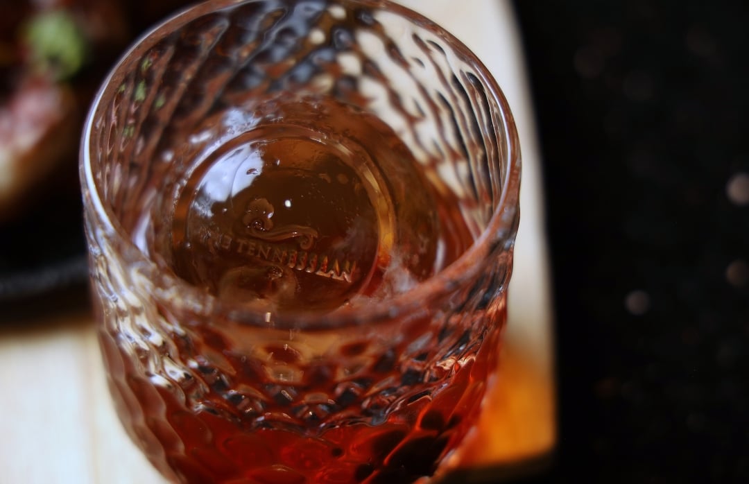 A close-up shot of a clear patterned glass holding an amber cocktail with a large round ice sphere.